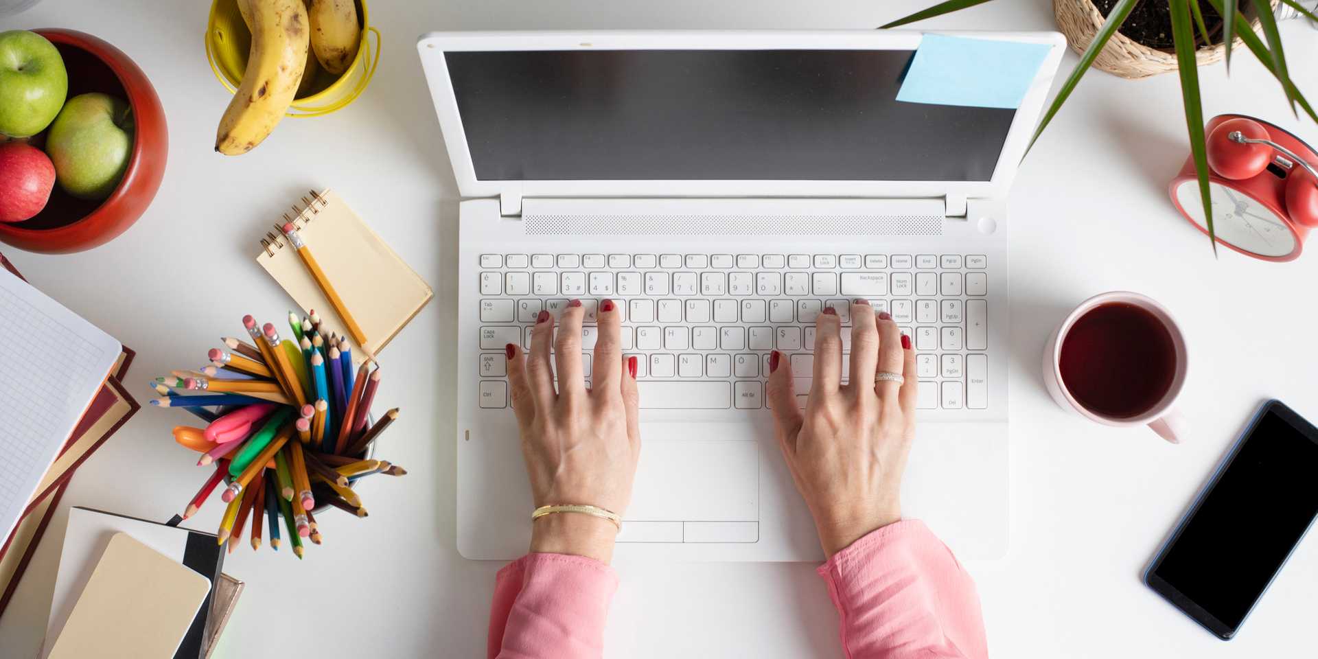 Overhead shot of woman working at laptop with fruit, pencils and smartphone on desk