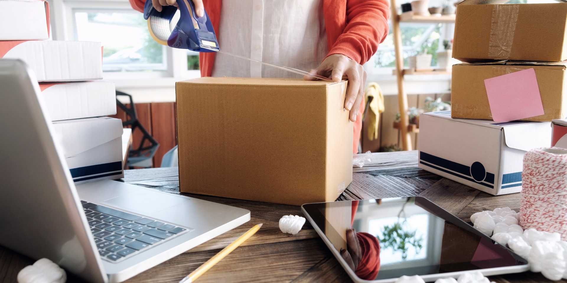 Person taping up a box in a shop surrounded by laptop, tablet and boxes